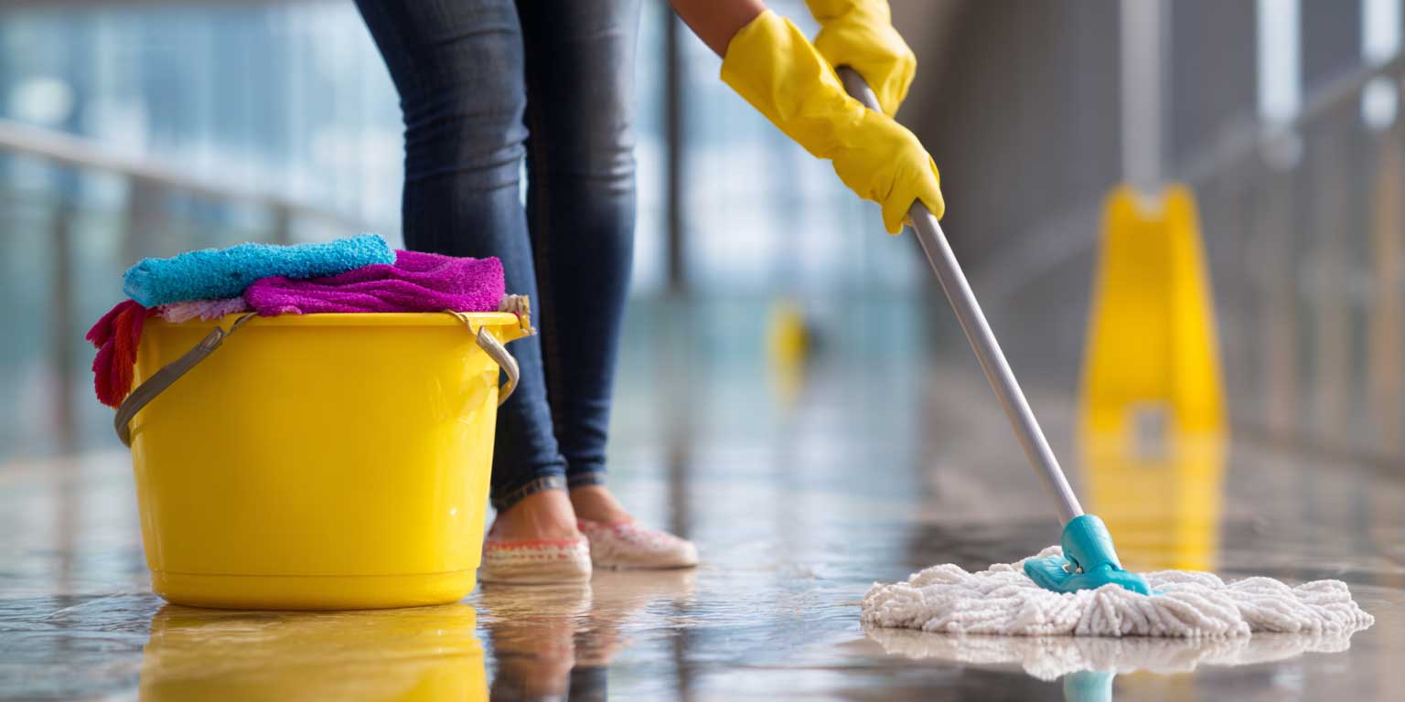 Yellow push broom sweeping a shiny wet tiled floor in a spacious room with large windows.
